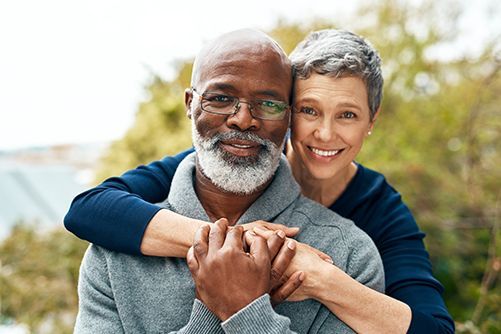 Two smiling adults hugging outdoors in a sunny park, with greenery blurred in the background