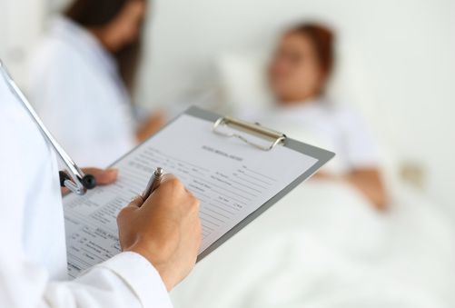Doctor writing on a clipboard beside a blurred patient in a bright hospital room