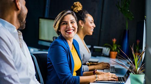 Colleagues smiling during a meeting at a conference table, with laptops and a plant nearby.