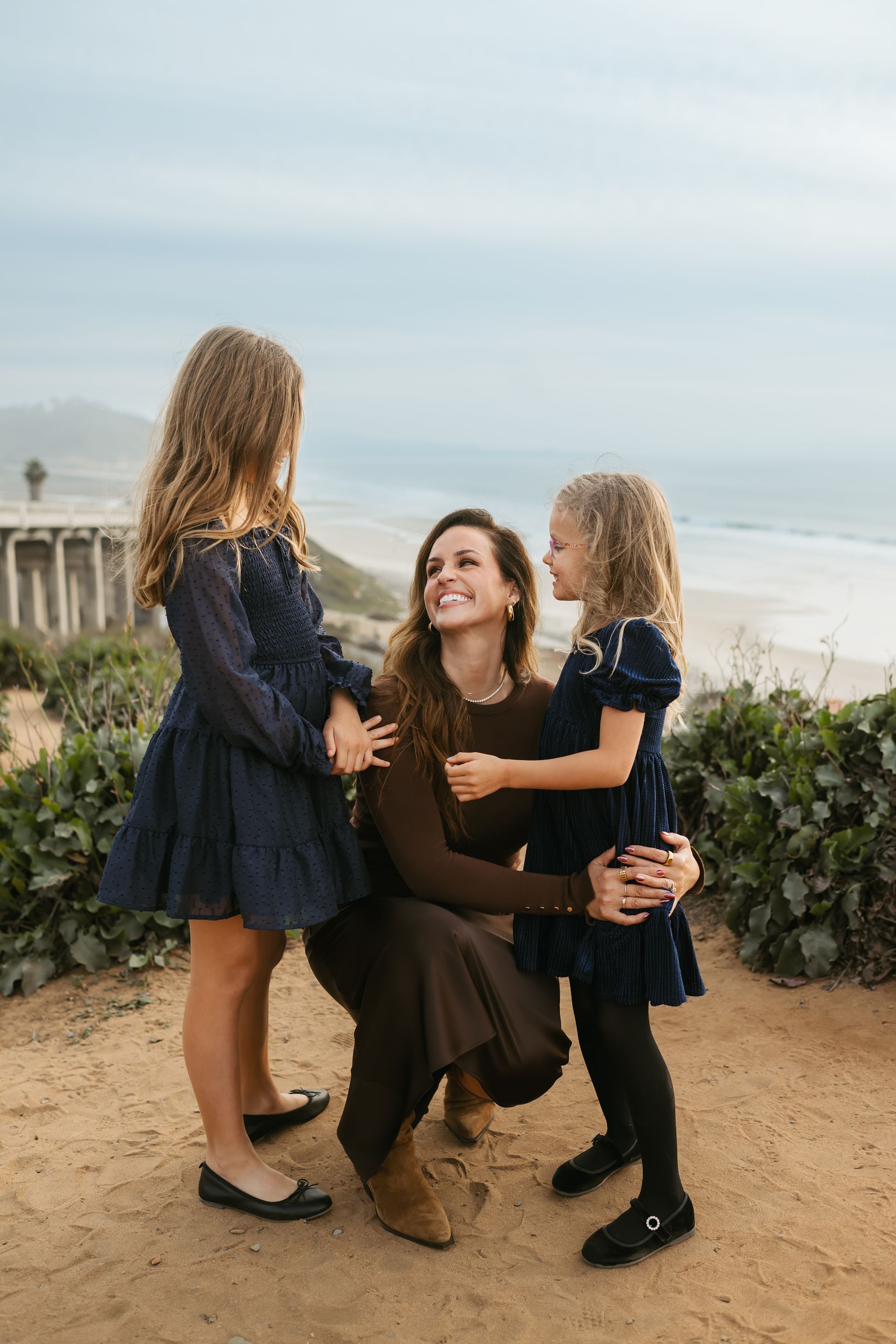 Woman kneels between two children on a beach, smiling and holding their hands.