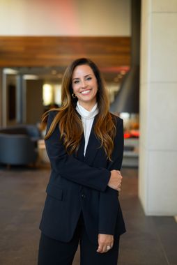 Smiling woman in a dark blazer standing indoors in a hallway with warm wood paneling and columns