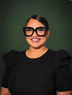 Woman in a black top and oversized glasses smiling against a dark green background