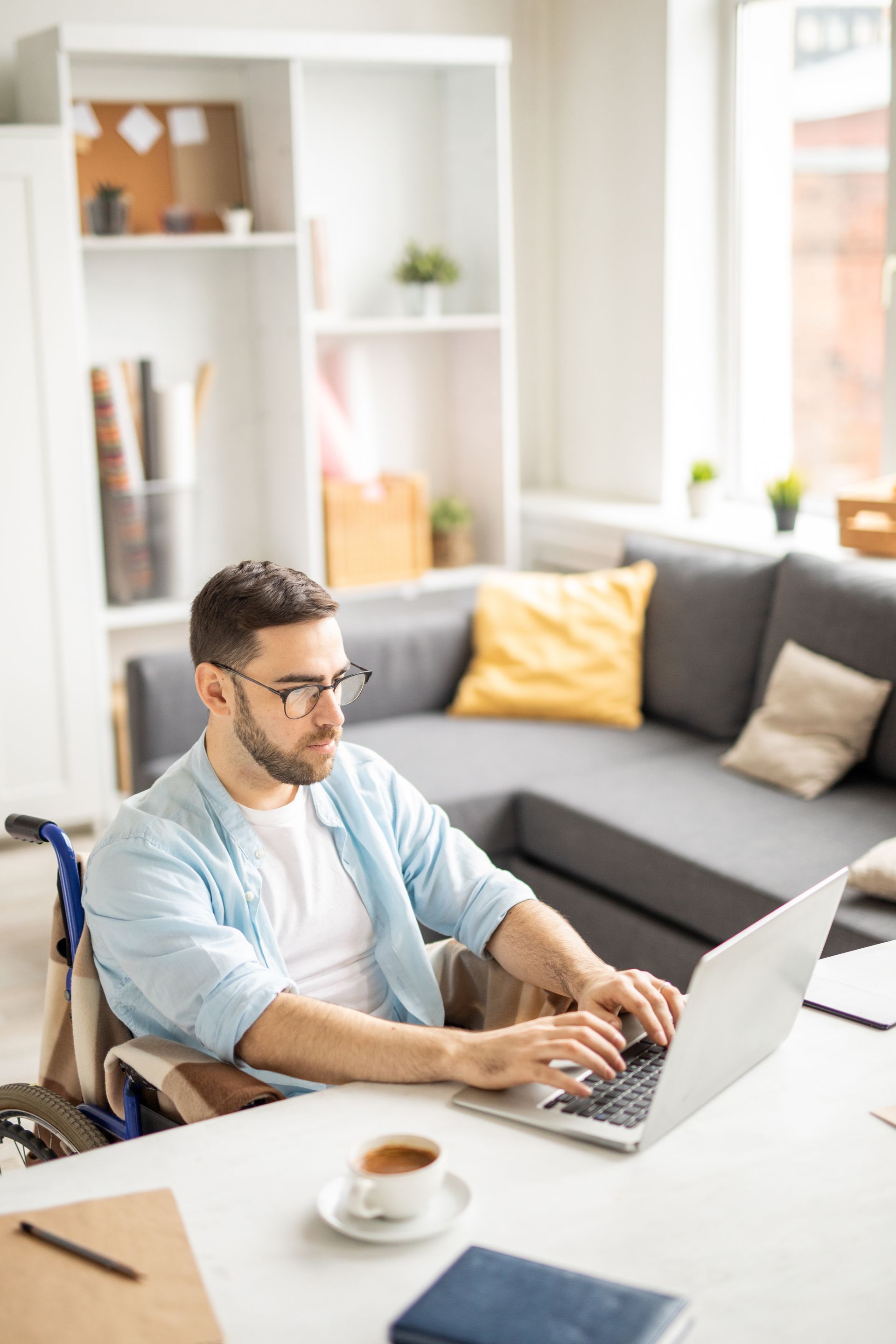 Man in a wheelchair typing on a laptop at a desk in a bright living room