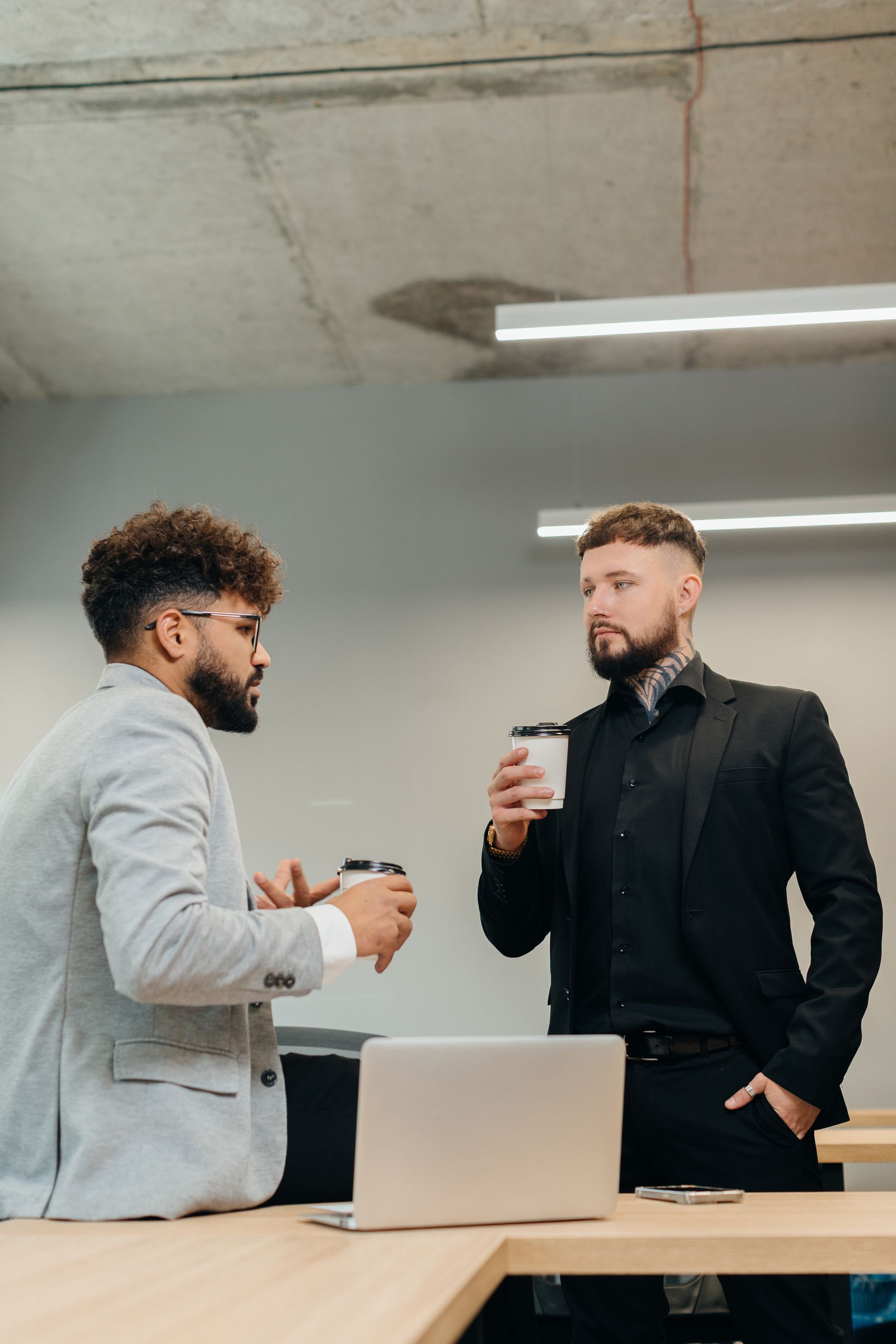 Two coworkers talking in an office, one holding a cup near a laptop on a desk.