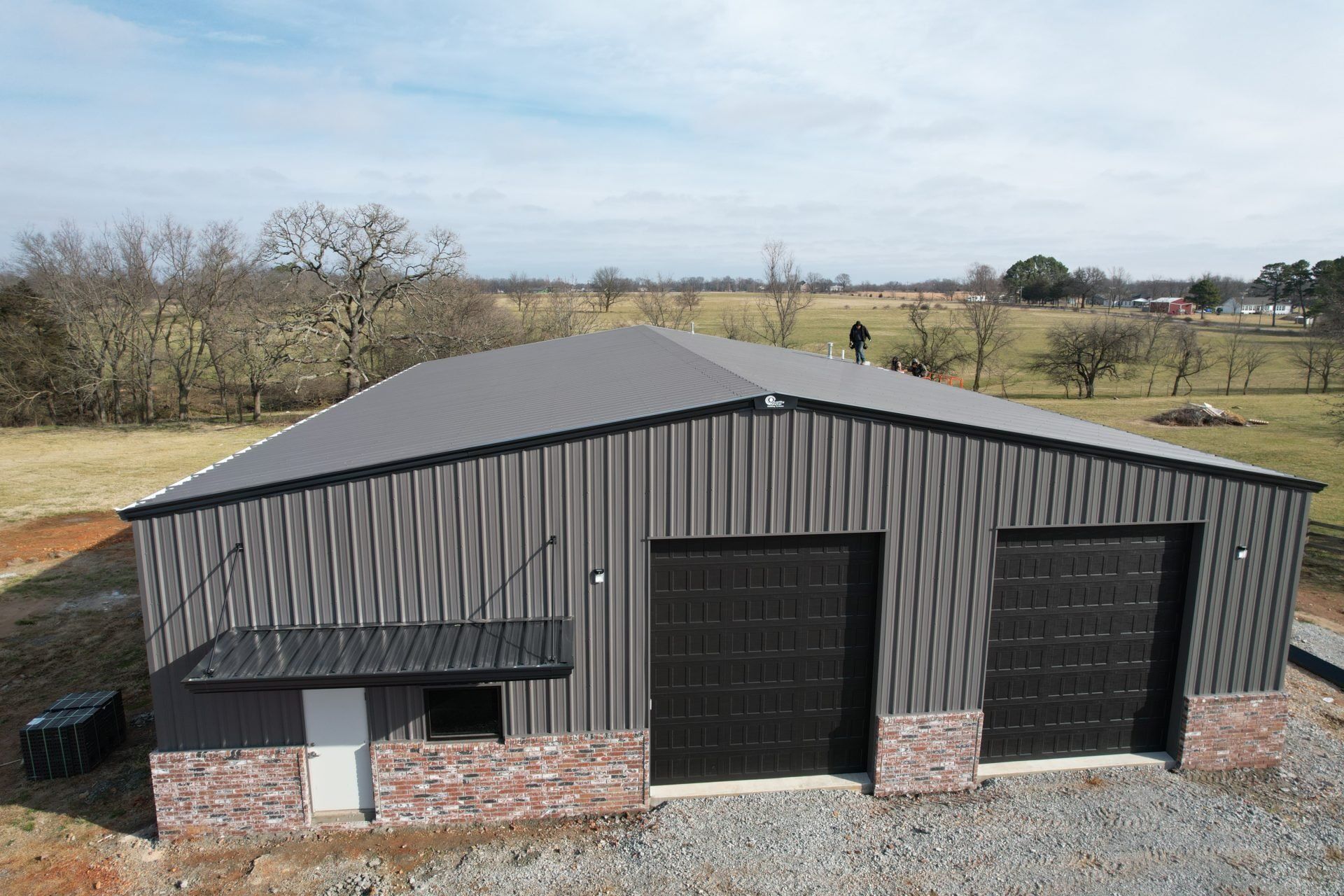 A gray metal-clad workshop with black garage doors and a brick facade sits in a rural setting under a clear sky.