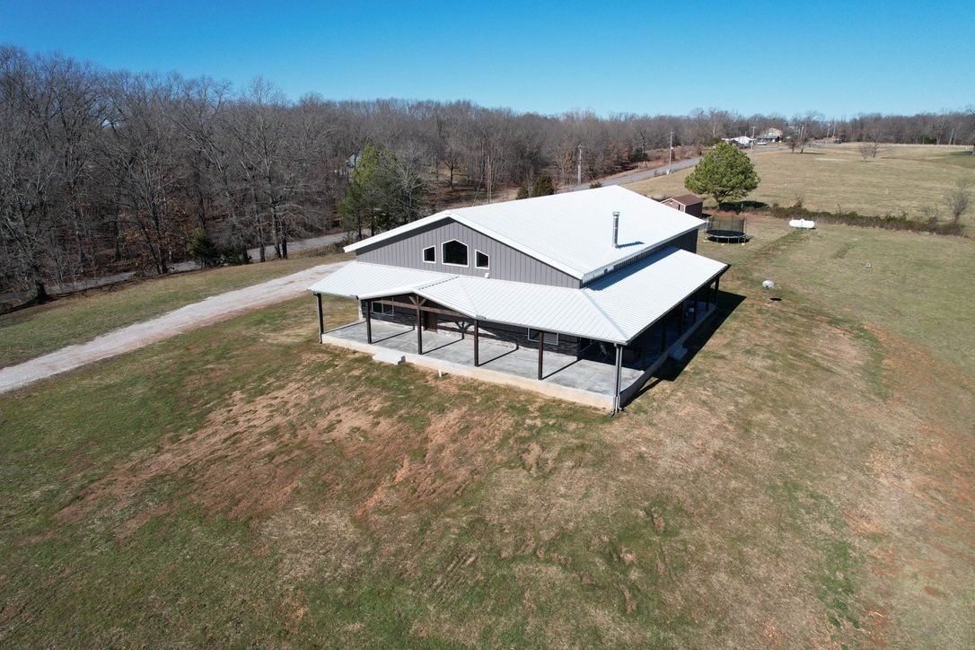Aerial view of gray metal home with white roof, covered porch, and gravel driveway in rural setting