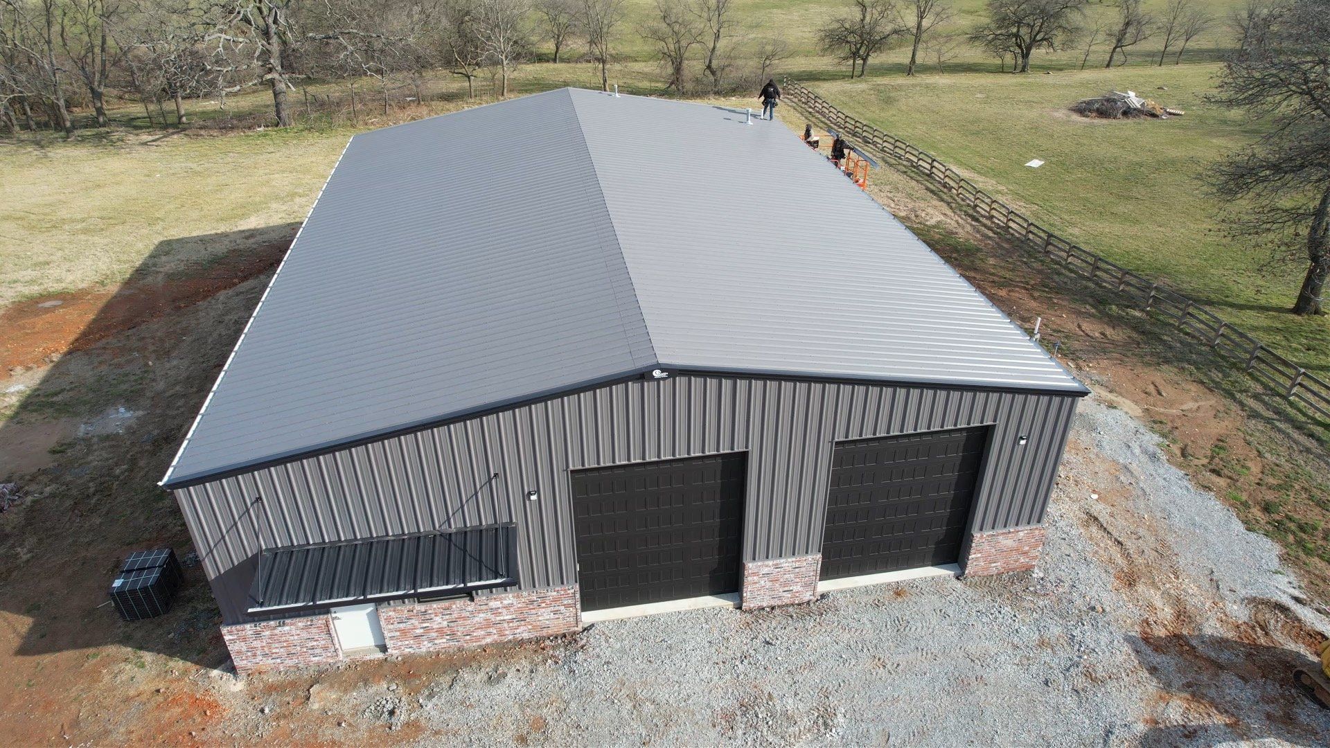 Aerial view of a gray metal building with a matching roof, two black garage doors, and a brick base in a rural setting.