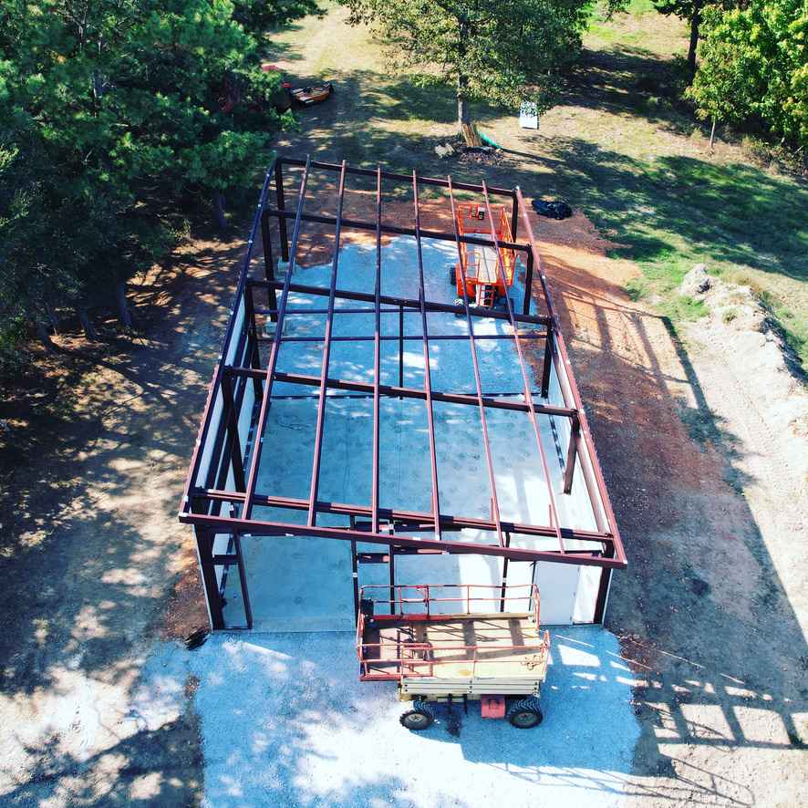 Aerial view of metal building frame under construction on concrete pad with lift and surrounding dirt and trees