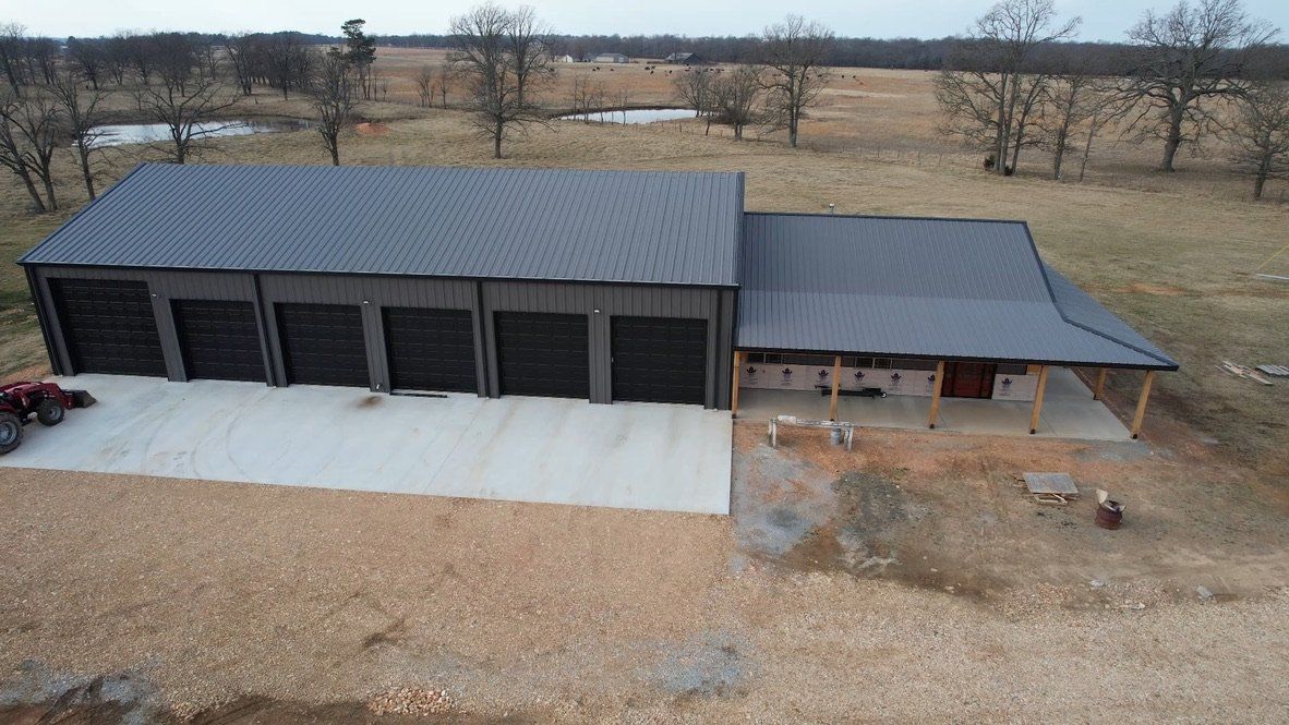 A large metal building with garage doors and a porch, in a rural setting, with a concrete pad in front.