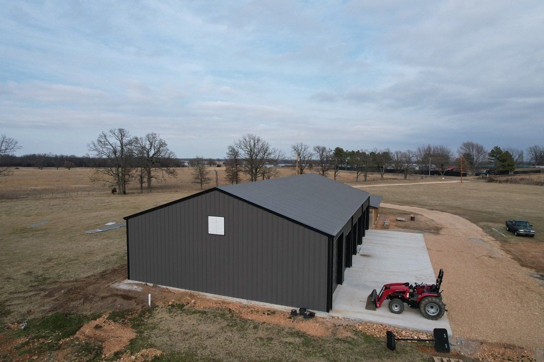 Dark gray metal building with garage doors, concrete pad, and tractor on gravel in rural setting