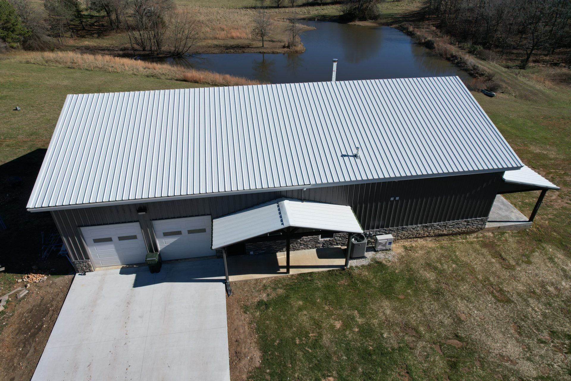 Aerial view of gray metal building with silver roof, garage doors, and concrete drive near pond