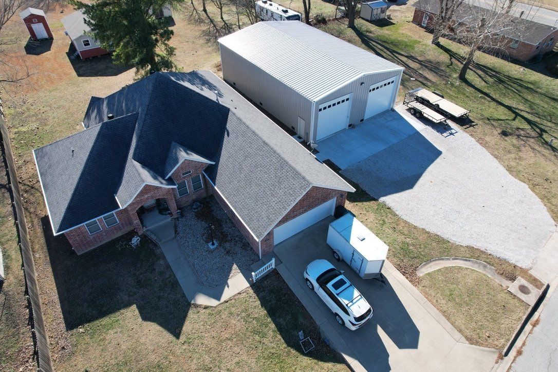 An aerial view of a brick house with a dark roof next to a large metal garage. A white car and trailer sit in the driveway.