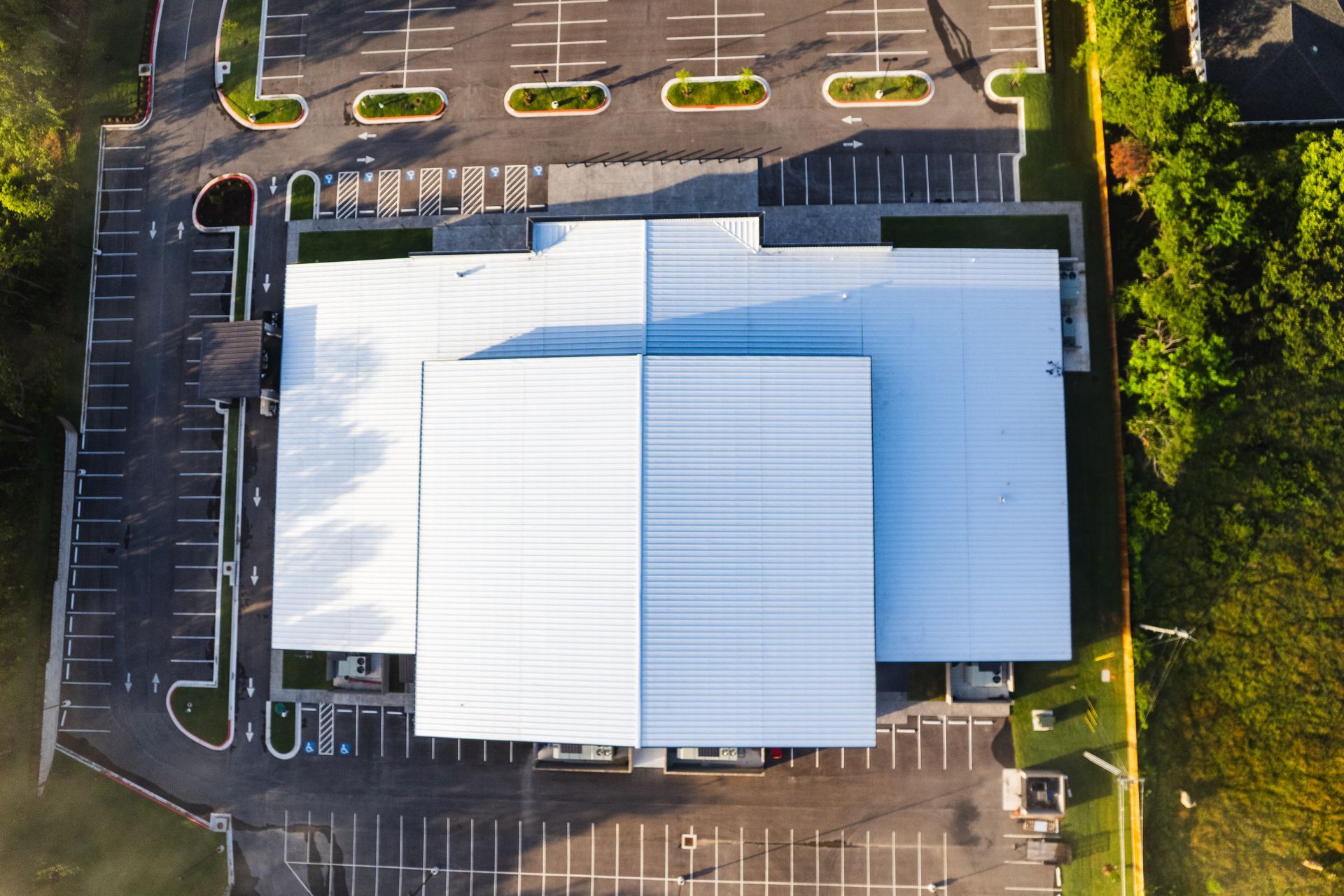 Aerial view of a large white-roofed building with a parking lot and landscaping; the building's entrance faces the parking.