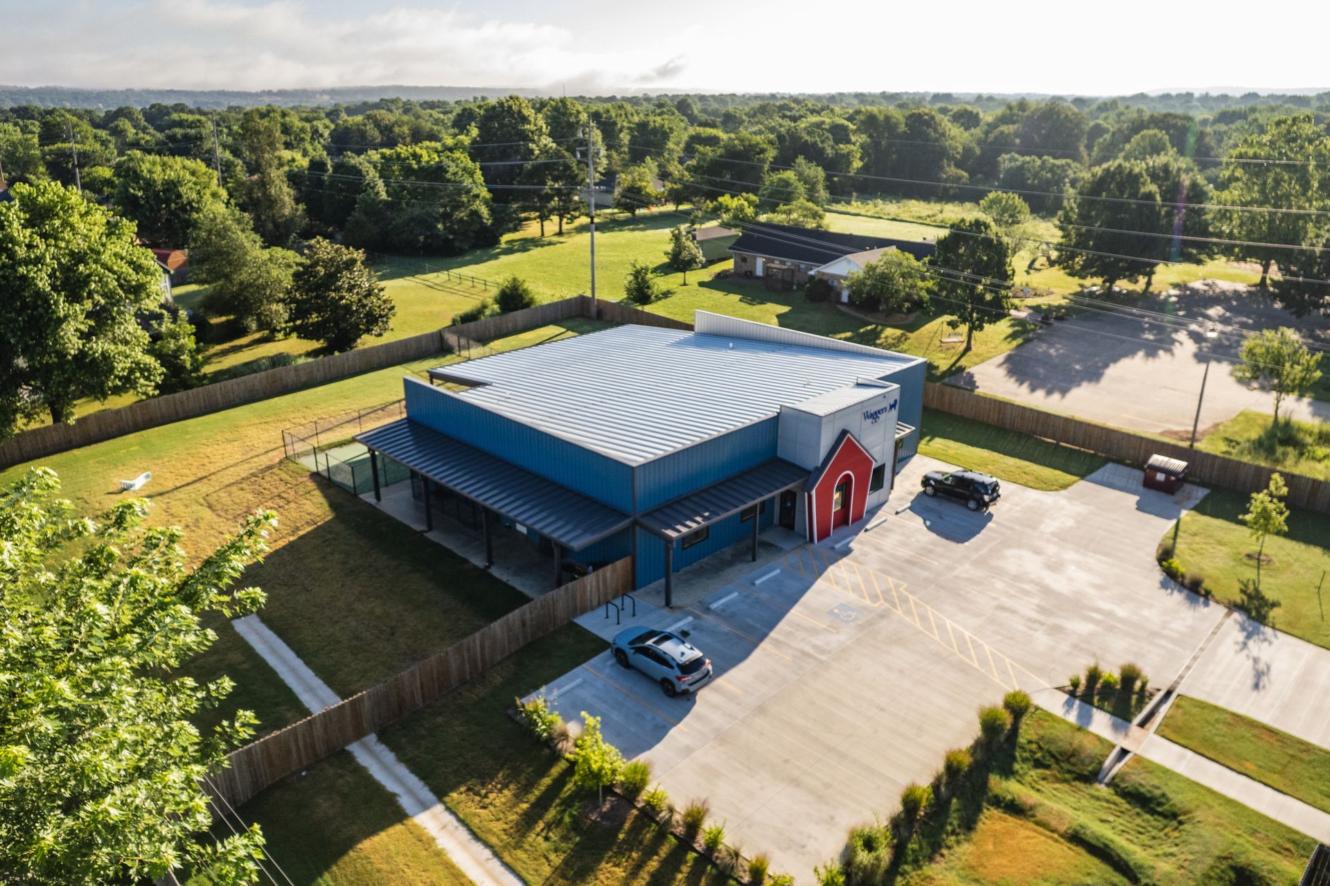 Aerial view of blue metal building with red entrance, corrugated roof, parking lot, and trees