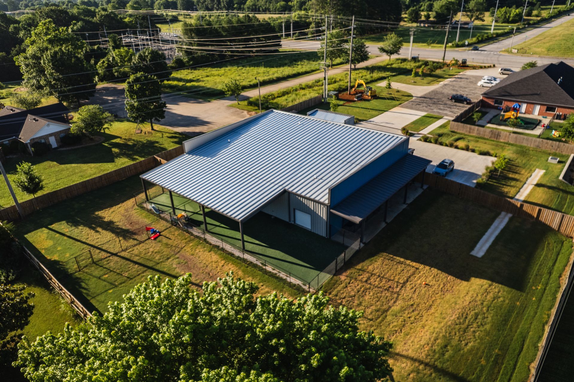 Aerial view of large metal building with corrugated roof on grassy lot with fence and nearby homes
