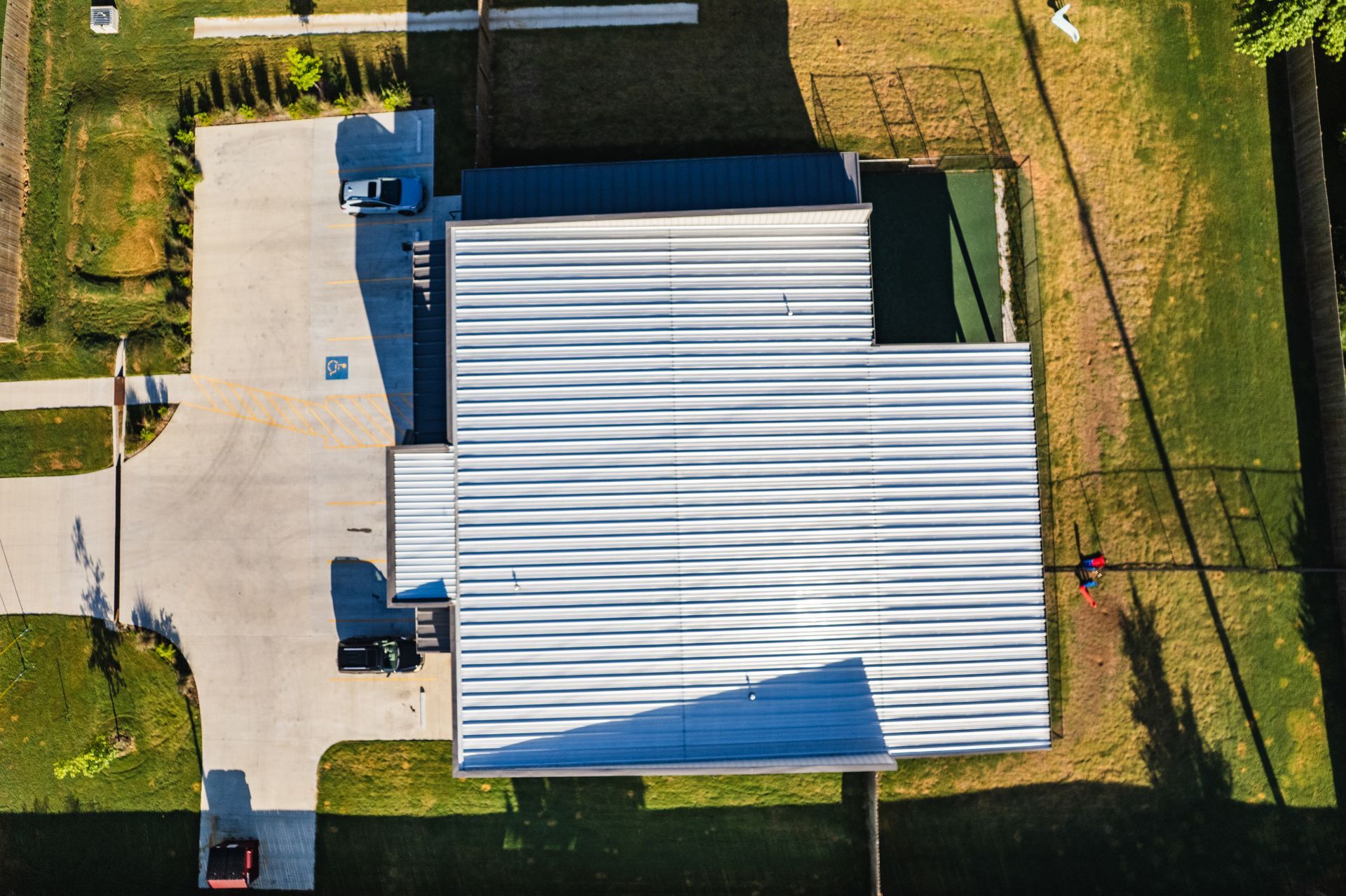 Aerial view of modern metal building with corrugated roof, driveway, vehicles, and green grass