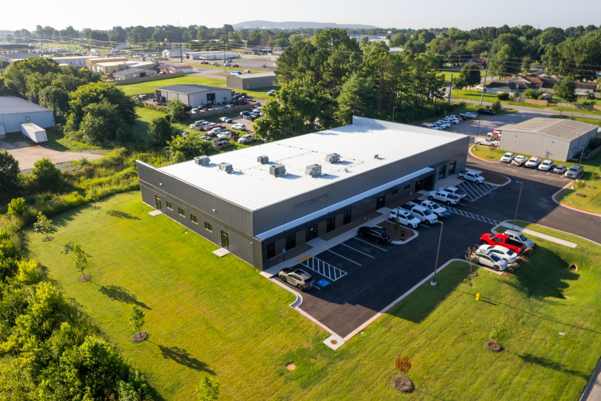 Aerial view of gray industrial metal building with white roof, parked cars, and trees on green lawn