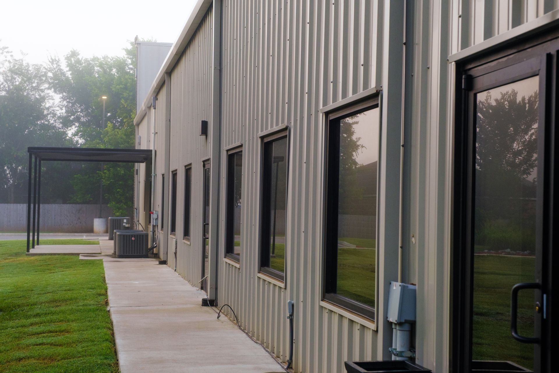 Corrugated metal building exterior with windows, glass door, covered walkway, and lawn on foggy day