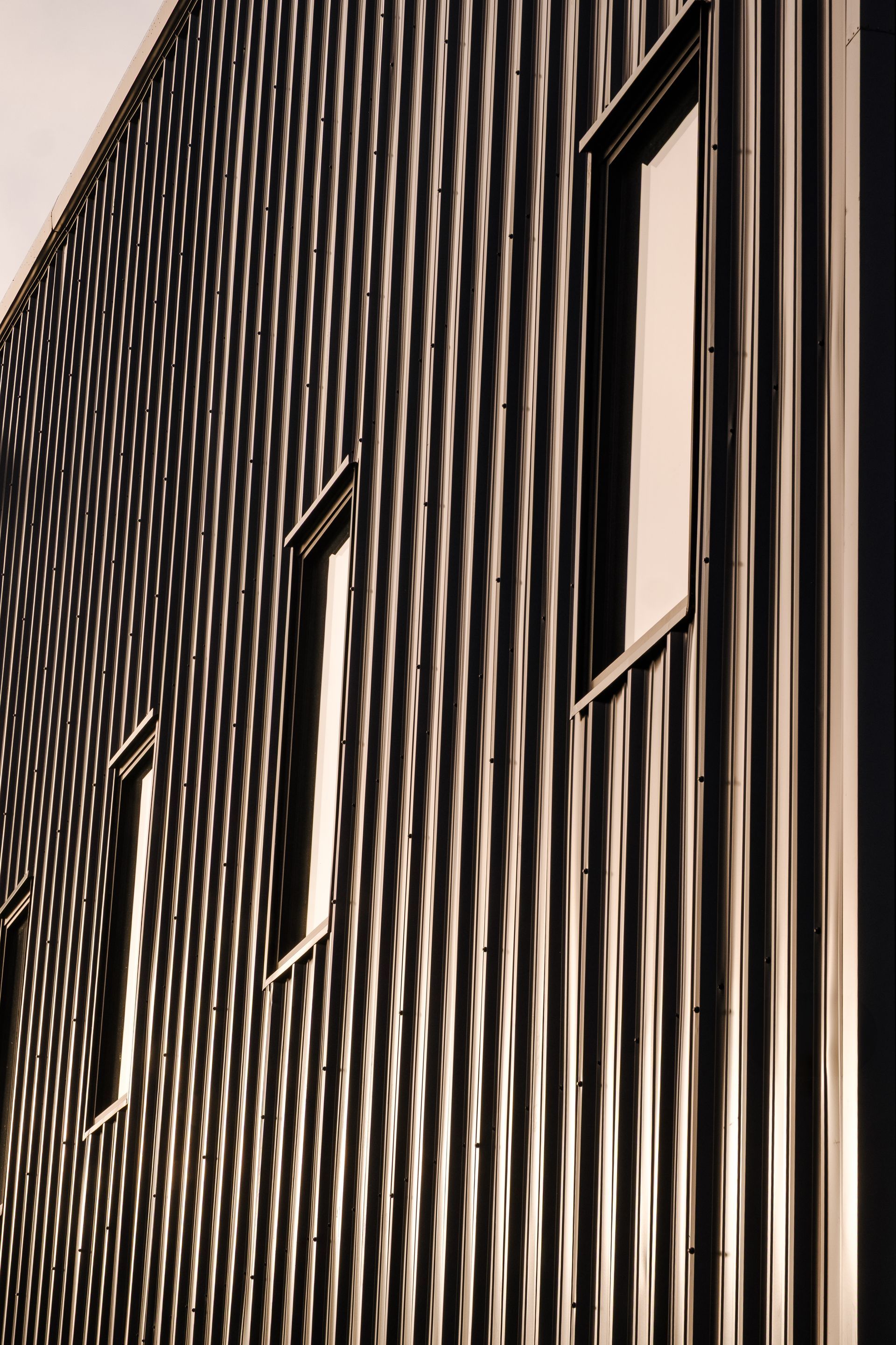 Side view of dark metal building with corrugated facade and three windows with sunlight shadows