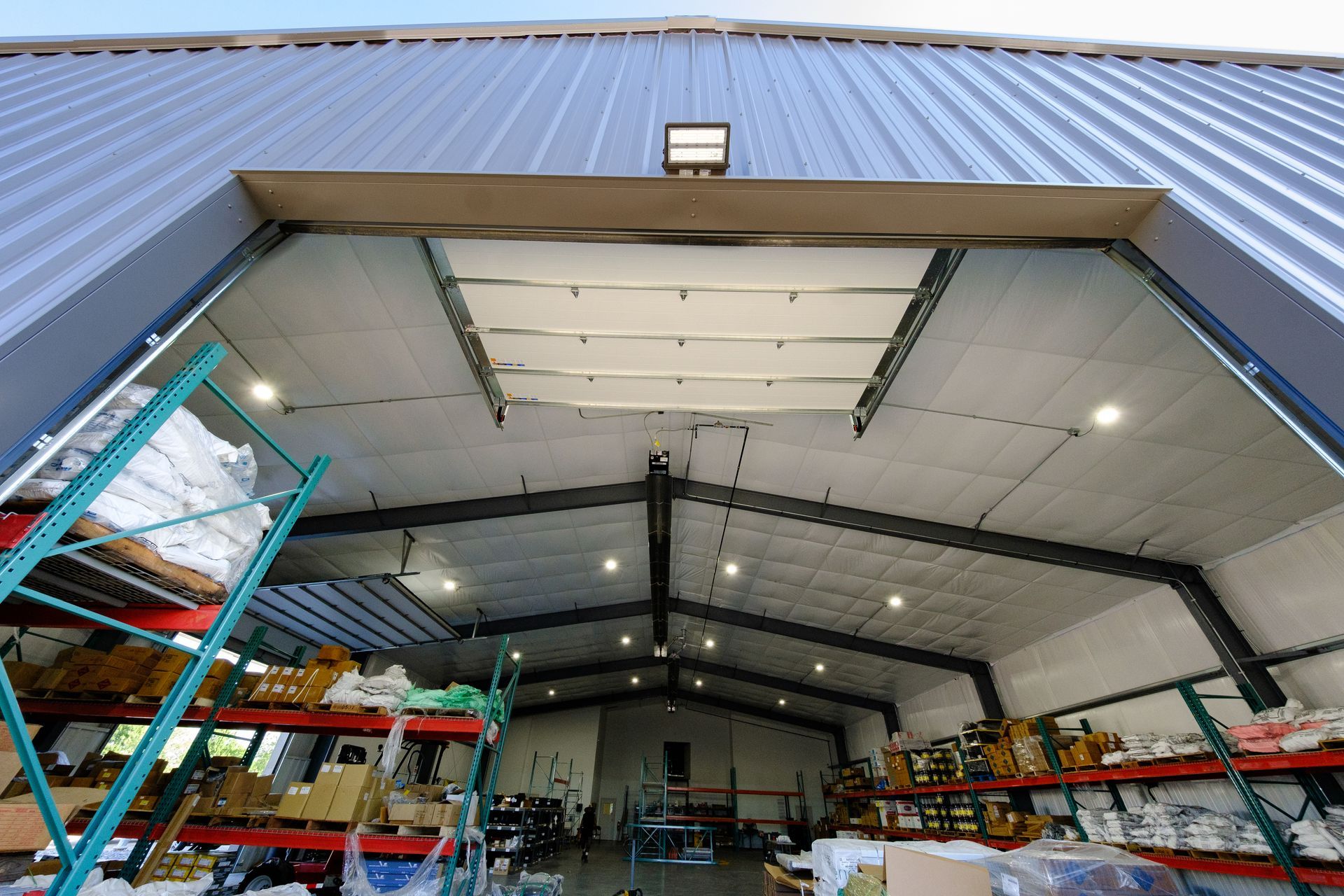 Warehouse interior with open overhead door, shelving rows, and bright lights from upward view