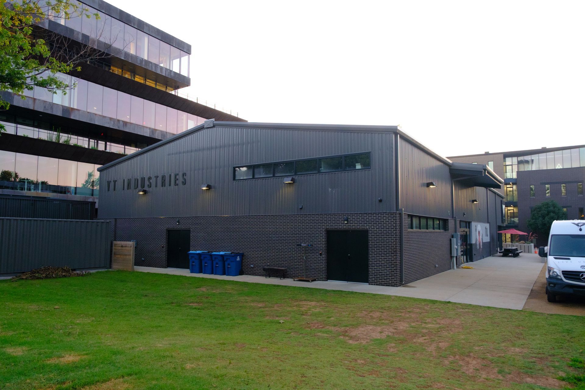 Dark gray corrugated metal building with PM Industries signage on grass with glass building behind