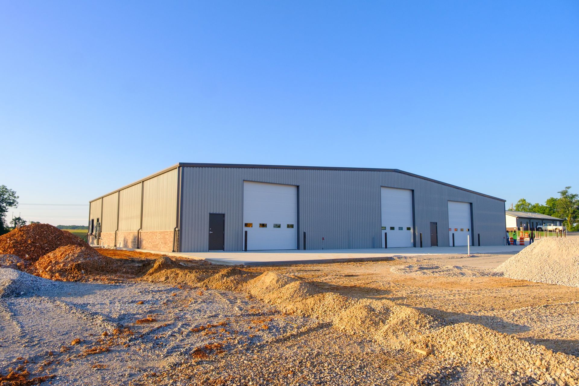 Gray metal building with garage doors on construction site with dirt piles under clear blue sky