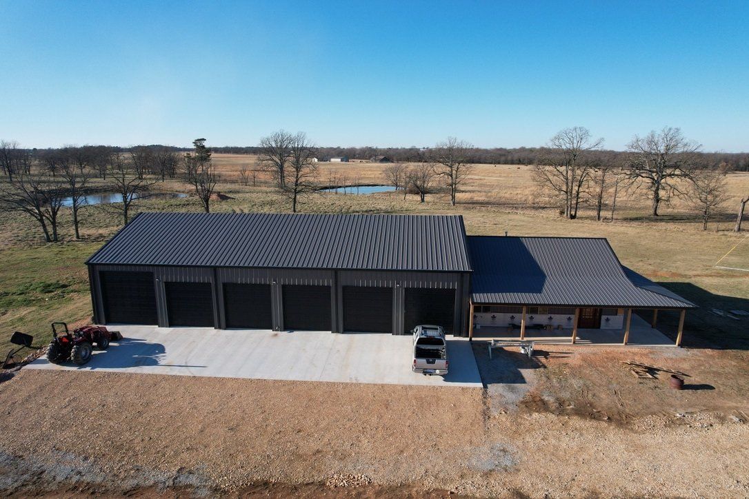 A large black metal-roofed building with garage doors and a covered porch in a rural setting under a blue sky.
