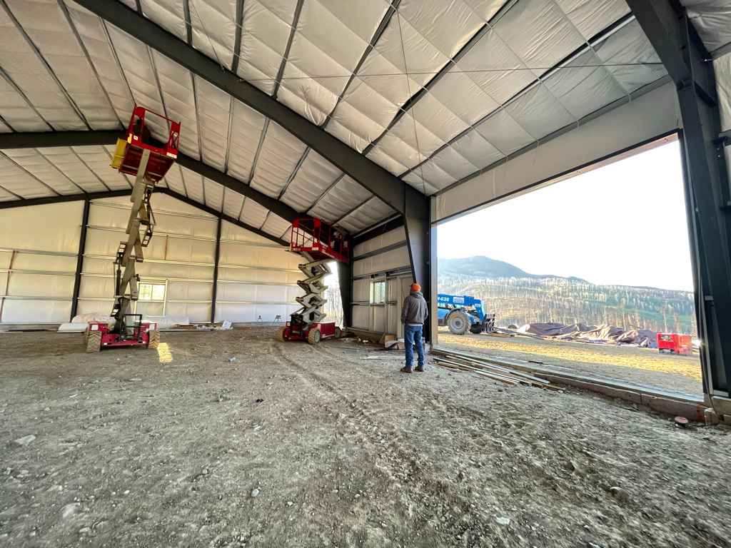 Man stands inside a new metal building. Two red lift machines are inside. The open doorway shows a mountain view.