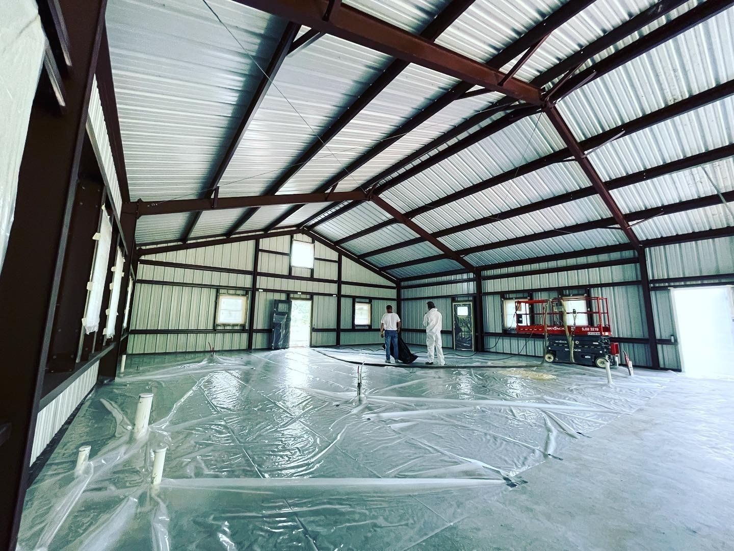Interior of a large metal building under construction. Workers are visible on the concrete floor covered in plastic sheeting.