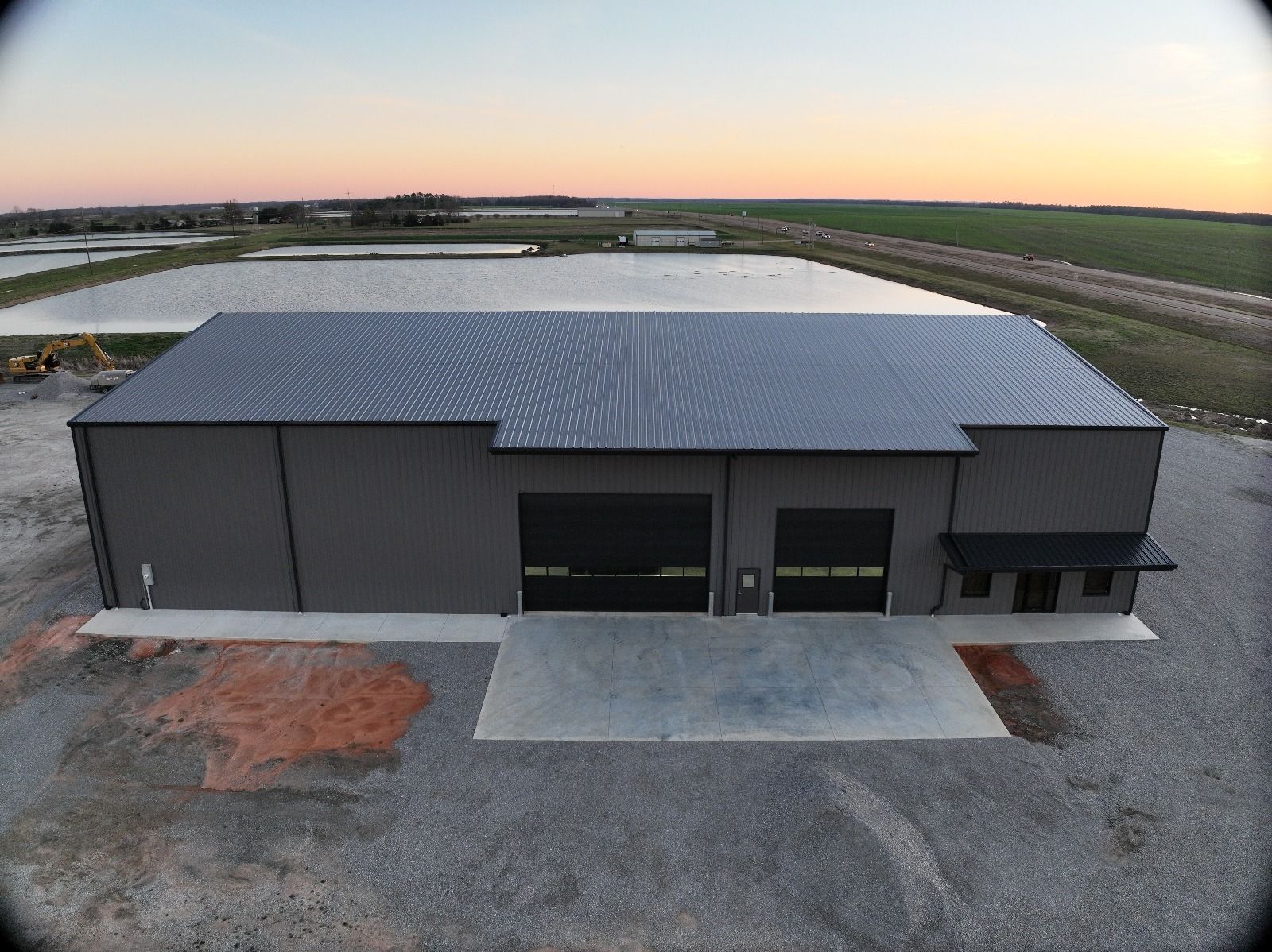 Gray industrial metal building with two garage doors and awning at sunset over ponds and fields