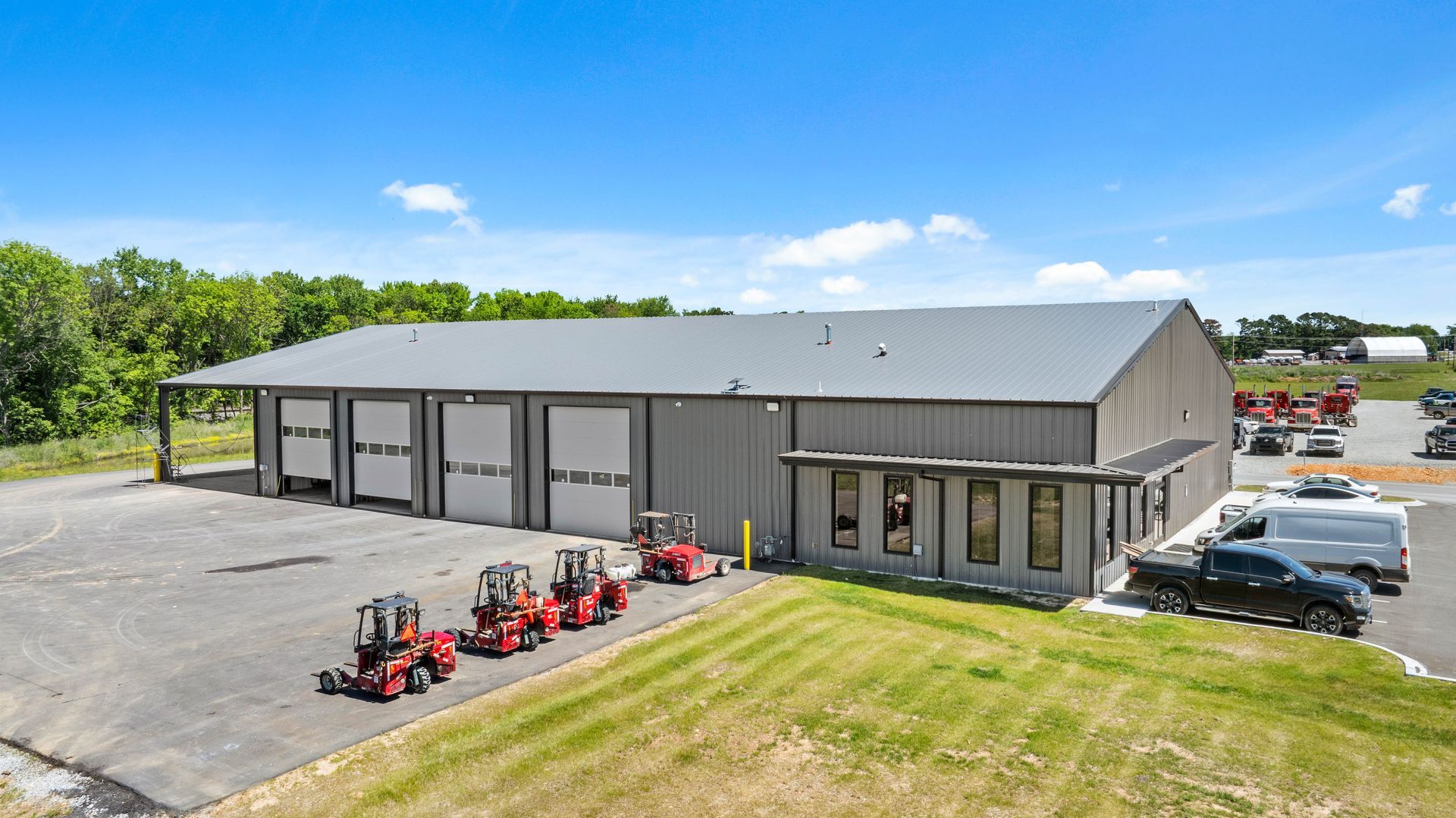 Gray industrial building with loading bays, parked trucks, and red lawn mowers in front on a sunny day