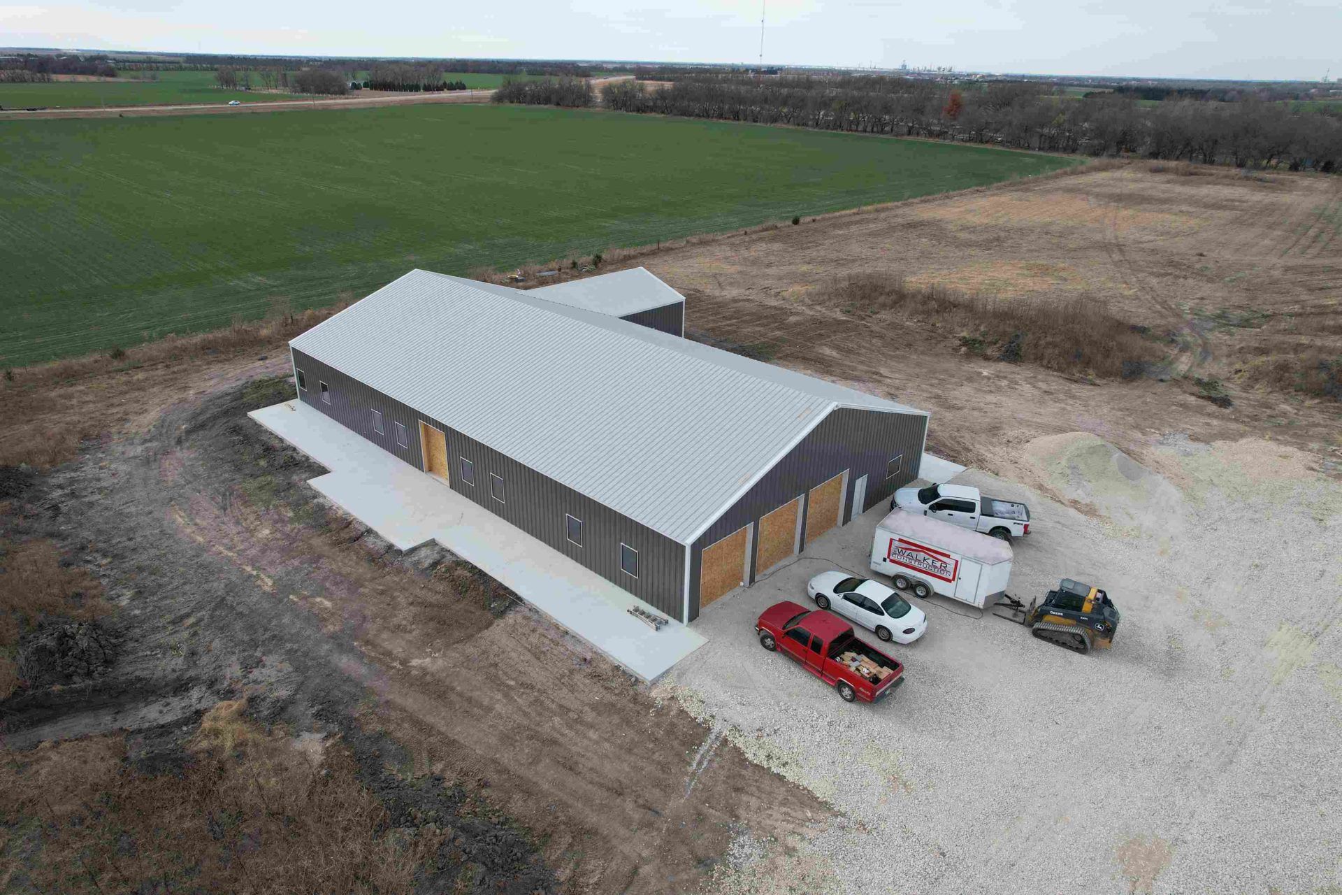 Aerial view of large gray metal building with silver roof, concrete pad, vehicles, and loader on gravel