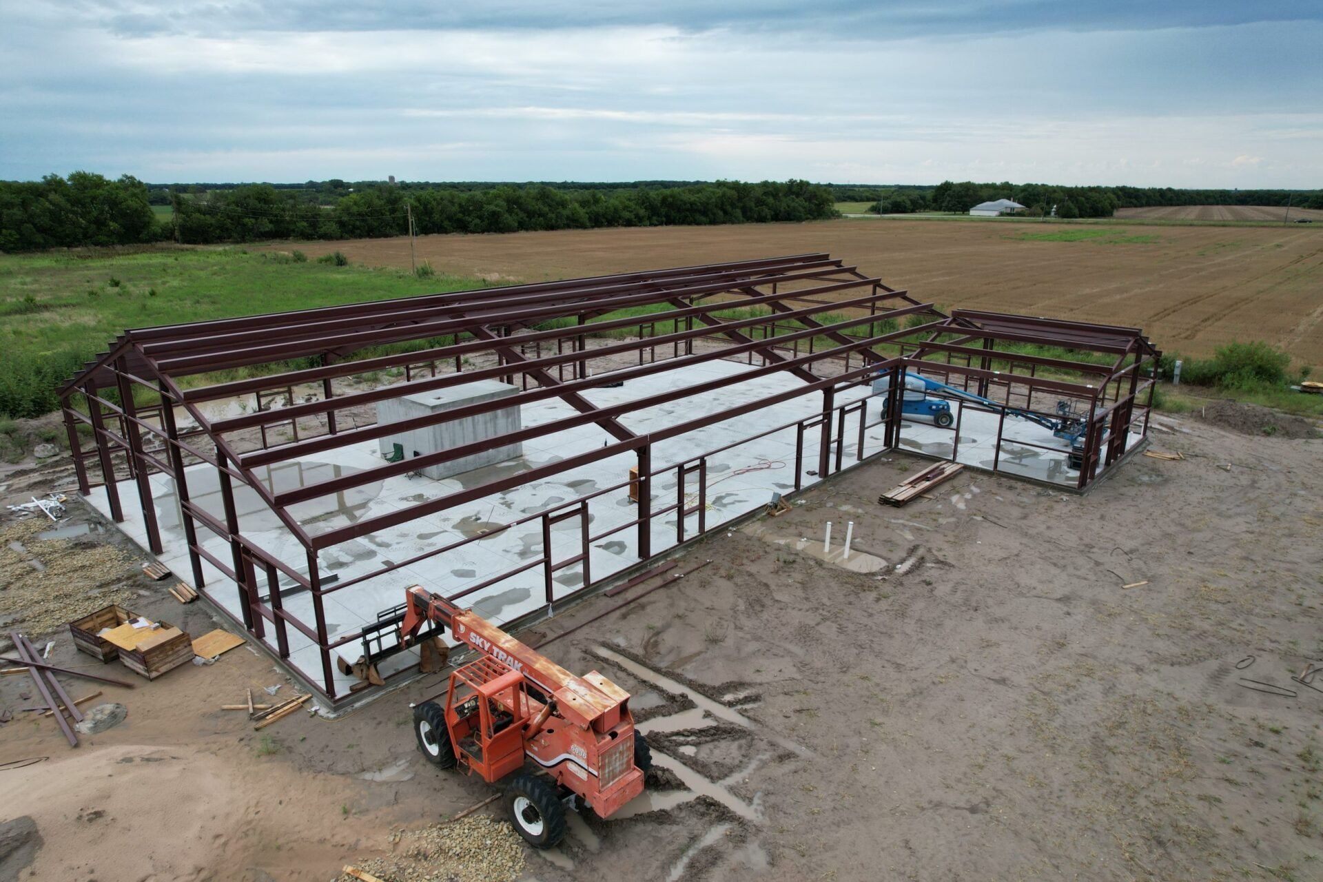 Aerial view of a steel-framed building under construction in a rural setting. A forklift is on the muddy ground.