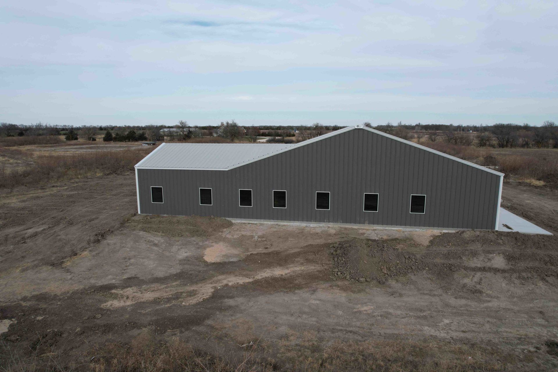 Gray metal building with silver roof and dark windows on dirt lot under cloudy sky