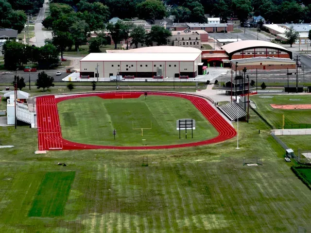 Aerial view of a red running track circling a grassy football field, adjacent to school buildings and spectator stands.
