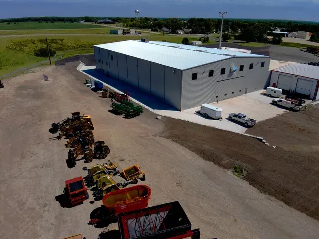 An aerial view of a large grey agricultural building with assorted agricultural equipment parked in a gravel lot nearby.