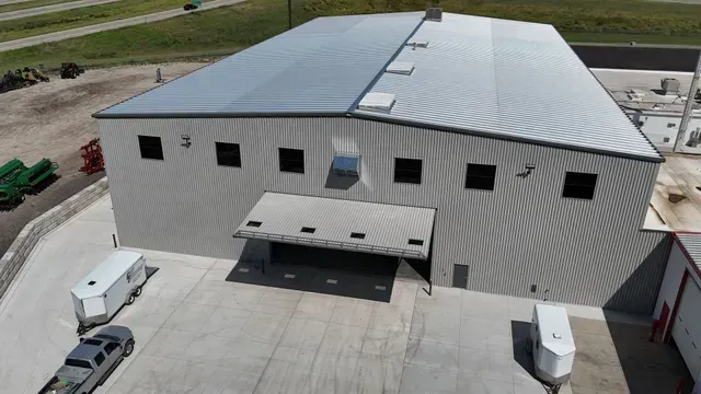 An aerial view of a large, gray industrial warehouse building with a metal roof and a loading dock area on a concrete lot.