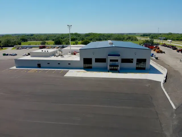 Aerial view of a gray, low-rise commercial metal building with a large paved parking lot on a sunny day.