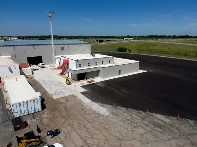 Aerial view of a white commercial metal building with a newly paved black asphalt section and construction equipment nearby.