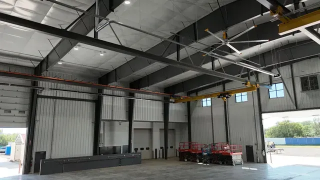 Wide shot of a large, empty industrial warehouse with high metal ceilings, steel beams, a crane, and a ceiling fan.