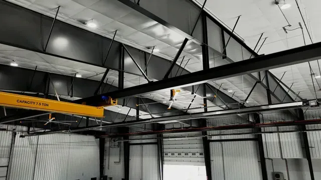 A yellow overhead crane bridge spans a high-ceilinged industrial facility with steel rafters and bright ceiling lights.