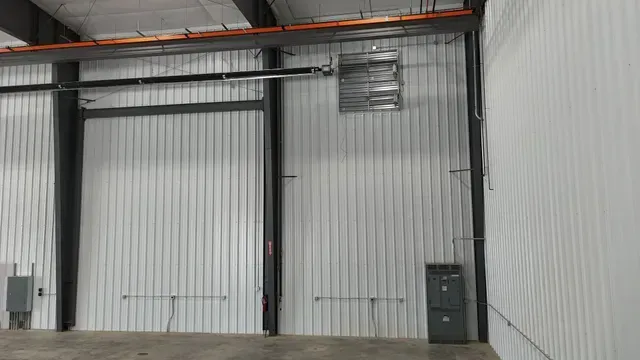 Interior of a metal building with white corrugated metal walls, steel beams, an overhead ventilation unit, and wall panels.