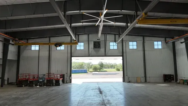 Inside a spacious industrial warehouse with white walls, a large open doorway, overhead crane rails, and a ceiling fan.