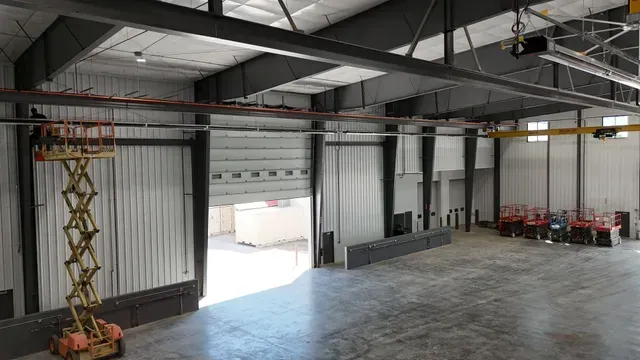 A large metal warehouse interior with a yellow scissor lift, exposed steel beams, high ceilings, and a wide open bay door.