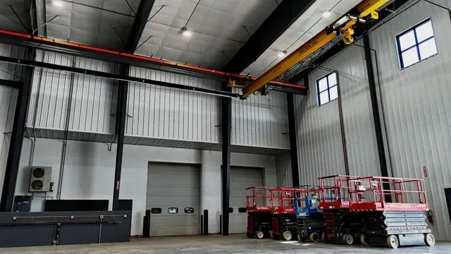 Industrial warehouse interior with a yellow overhead crane, two large roll-up doors, and several red scissor lifts.