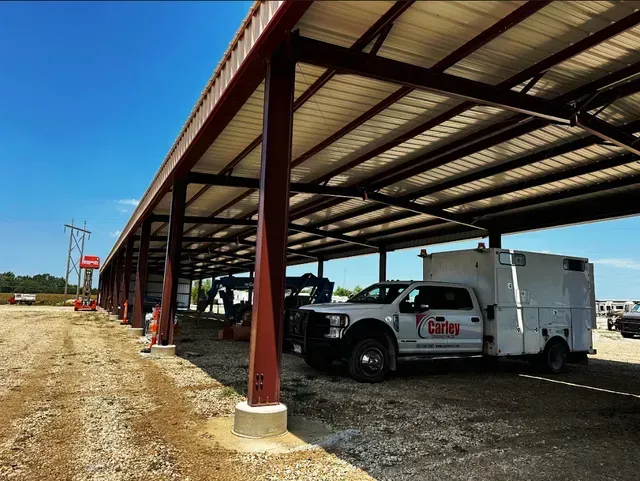 A white utility truck parked under a large, open-air steel canopy on a gravel lot under a clear blue sky.