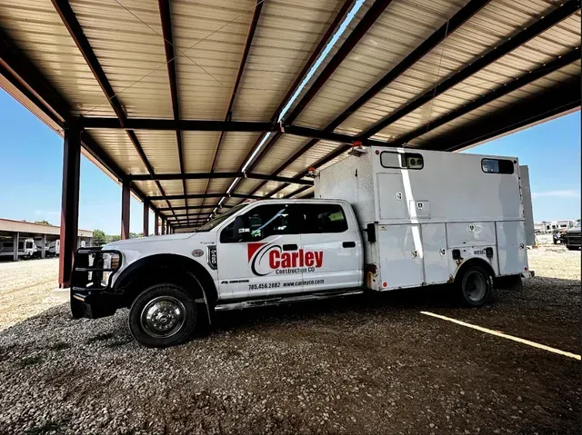 A white service truck with the Carley logo parked under a covered metal structure on a gravel lot.
