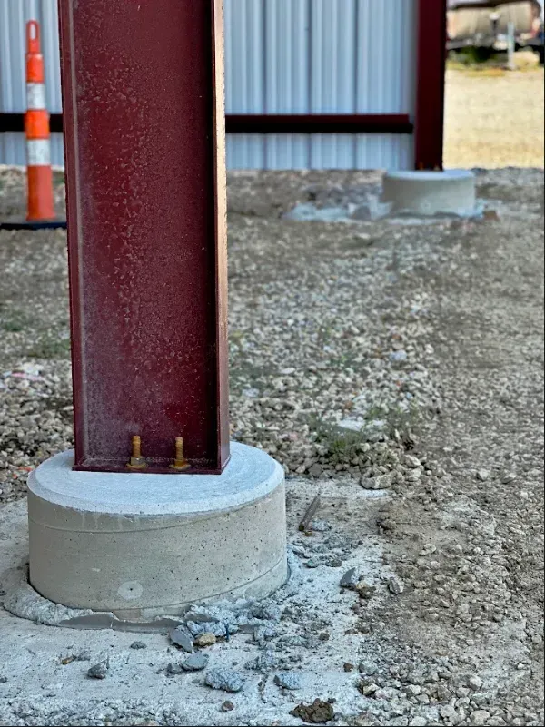 A red metal I-beam column mounted on a circular concrete pier, set in a gravel area with a metal building wall behind.