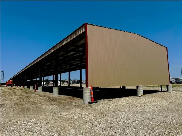 A long, open-sided steel pole barn with tan metal siding and red framing, situated on a gravel lot under a blue sky.