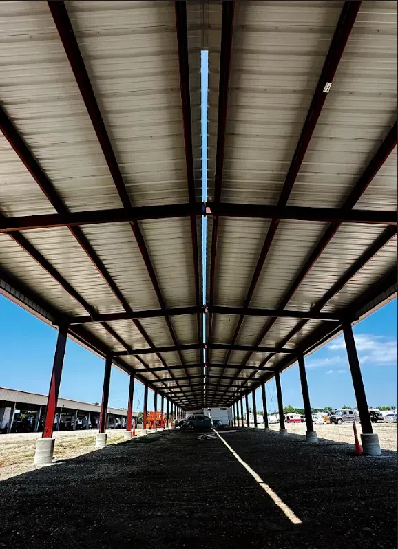 An outdoor metal carport shelter with a central open ridge vent, steel beams, and a gravel parking surface.